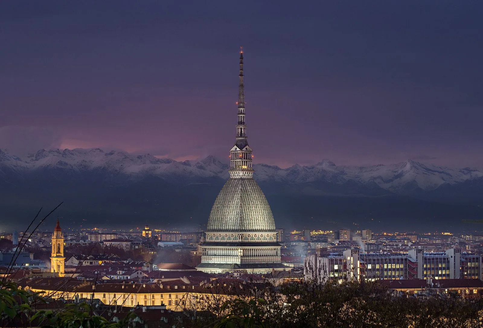 La Mole Antonelliana illuminata al crepuscolo, con i tetti di Torino in primo piano e le Alpi innevate sullo sfondo