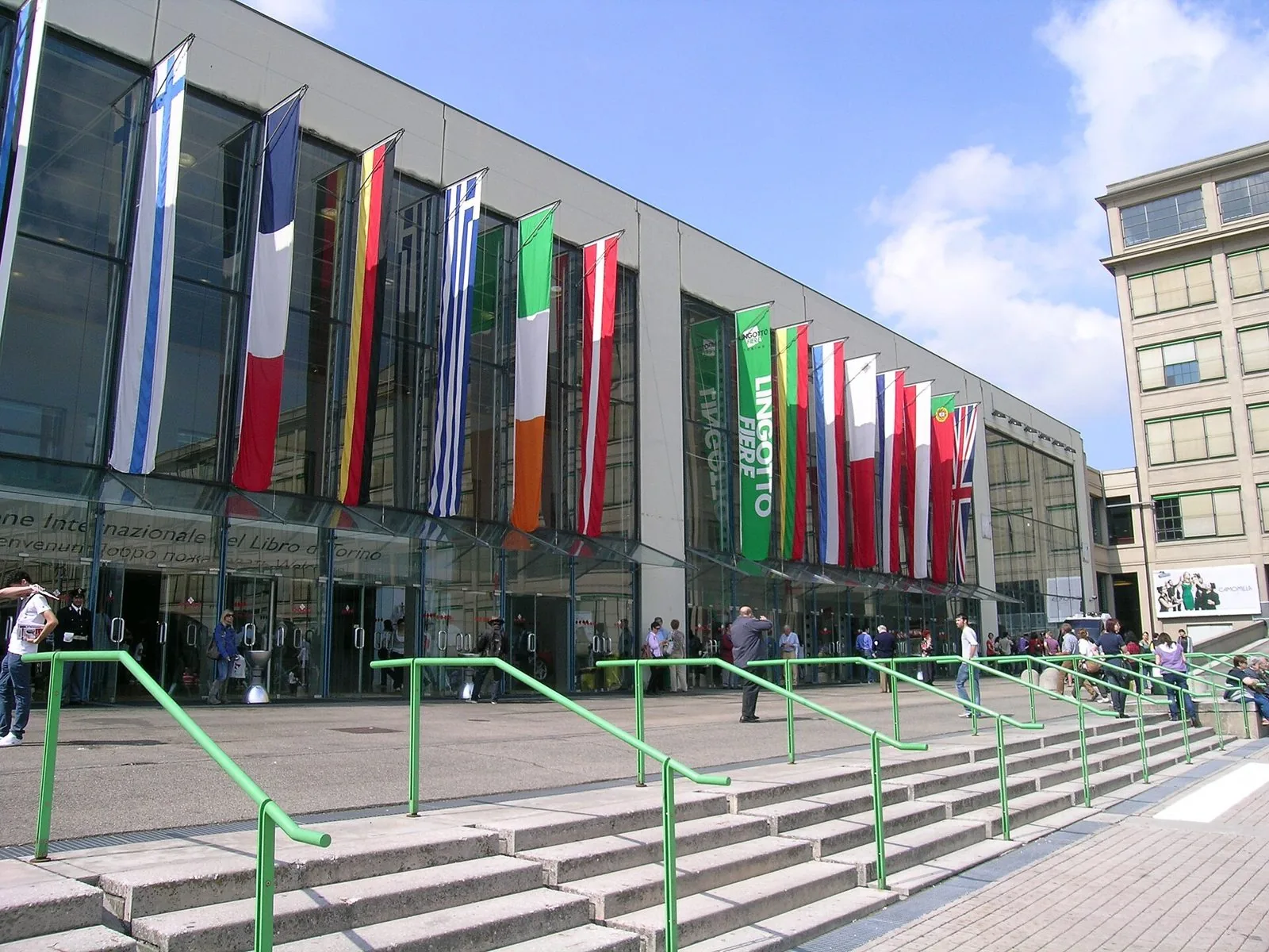 L'ingresso di Lingotto Fiere a Torino durante il Salone del Libro, con le bandiere di vari paesi appese alla facciata vetrata e la scalinata con corrimano verdi